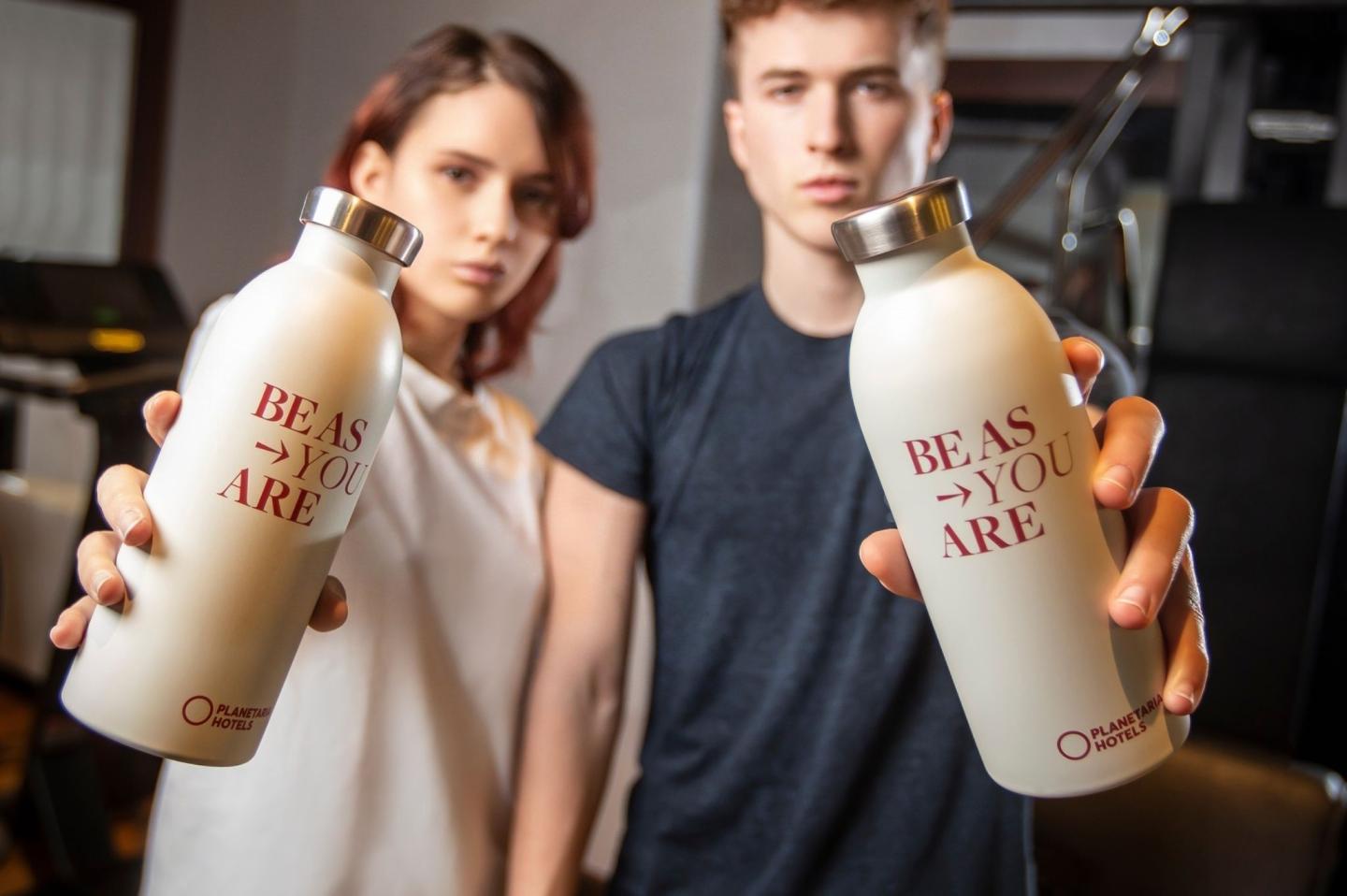 Two people holding up cream-colored water bottles with the red text BE AS YOU ARE and the Planetaria Hotels logo